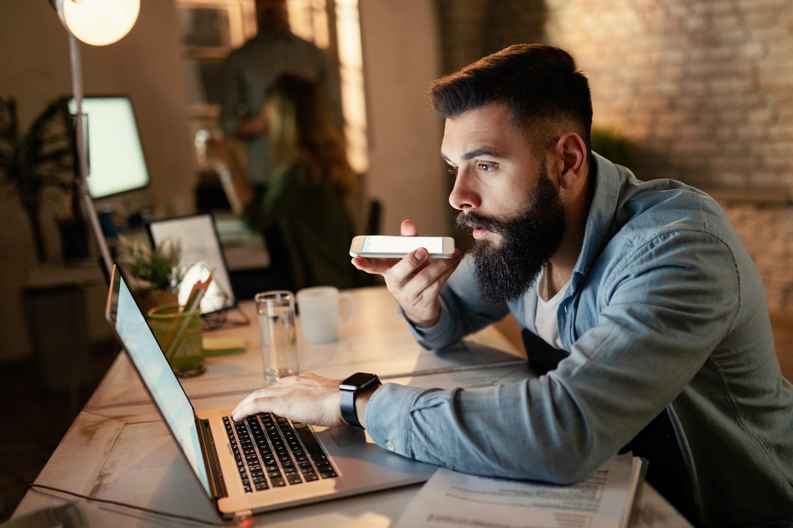 Young entrepreneur reading an e-mail on computer while recording voice message on smart phone during late night work in the office.